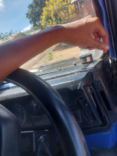 The scene is set inside a vehicle in Kimamba, Tanzania, observed from a forward-looking perspective. A right human arm with medium skin pigmentation is visible, extended towards the upper right, with the hand's fingers positioned near the windshield frame or A-pillar. A black steering wheel occupies the lower-left foreground. The dashboard, dark in color, spans the lower half of the frame, displaying part of an instrument cluster with a fuel gauge indicating a half-full reading. A small, metallic, cylindrical object rests on the dashboard near the windshield. Through the windshield, a bright blue sky with minimal cloud cover is visible, along with a large tree bearing yellow blossoms in the upper right. A dirt road and distant single-story buildings with corrugated roofs are present in the background. The environment is under bright daylight.