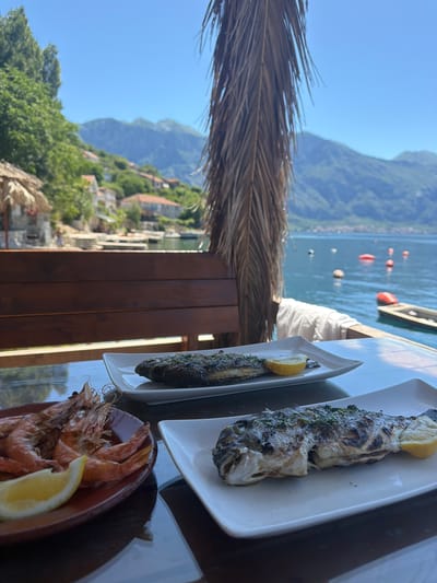 The image captures a meal at a restaurant in Dražin Vrt, Montenegro, overlooking the Bay of Kotor. The foreground features a wooden table laden with seafood dishes: grilled fish on white plates, each garnished with a lemon wedge, and a terracotta bowl filled with shrimp, also with a lemon wedge. A dried palm frond partially obscures the view on the left. The background reveals a picturesque scene of Dražin Vrt, a village nestled along the bay's edge, with buildings scattered among lush greenery leading up to the mountains. The clear blue sky contrasts with the calm water of the bay, dotted with buoys and boats.