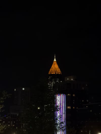 The image depicts a night scene in Atlanta, United States of America. The Georgia World Congress Center is identifiable by its illuminated signage. In the background, a tower with its spire is visible in yellow lights. Trees are in the foreground. The image is oriented upside down.
