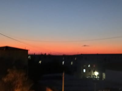 A dusk scene captures the sky over Qorowul, Uzbekistan, transitioning from a soft blue to a vibrant orange gradient. Silhouetted against the colorful sky are the dark forms of buildings, some with illuminated windows. A faint wire traverses the sky, and a small, indistinct cloud is visible in the upper right quadrant. The foreground is dominated by the dark shapes of structures, with hints of foliage in the lower left. The overall impression is one of a quiet urban evening.