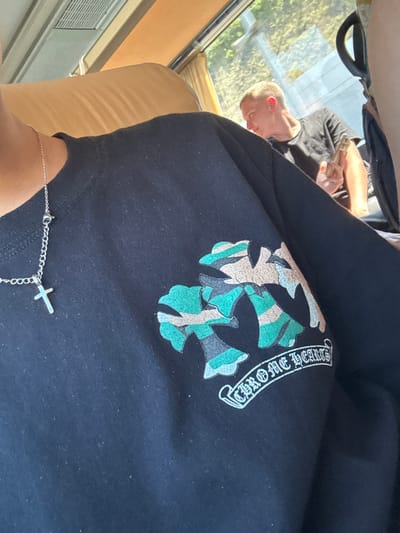 The image shows a person lying down on a beige cushioned surface, possibly a couch or a car seat, with their head resting on their arm. They are wearing a black t-shirt with a camouflage-style cross graphic and the text "CHROME HEARTS" emblazoned on it in white lettering. A delicate silver necklace with a small cross pendant is visible on their wrist. In the background, a window reveals a sunlit outdoor scene with trees and foliage, suggesting it is daytime and likely pleasant weather. The lighting indicates a bright, sunny day. The overall setting appears to be a relaxed, indoor environment with a view of nature.