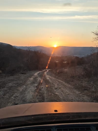 The image displays a sunset over the city of Opaka, Bulgaria, captured from the interior of a vehicle. A winding, unpaved dirt road with visible ruts and muddy patches dominates the lower-middle section of the frame, receding into the distance. The sun, a bright orange disc, is positioned low on the horizon, partially obscured by rolling hills. A distinct vertical lens flare extends downwards from the sun onto the road and the vehicle's hood. The sky above transitions from a vibrant orange at the horizon to a pale blue with scattered clouds higher up. The surrounding landscape consists of denuded trees and dry, sparse vegetation, indicating a colder season. In the distant valley below the sun, structures consistent with a town or city, identified as Opaka, are visible. The top edge of a dark vehicle dashboard and the front portion of its hood are present in the immediate foreground, reflecting the orange light. A small, dark, unidentified object is airborne in the sky to the left of the sun.