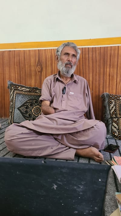 An elderly man with gray hair and a beard is sitting on a patterned cushion in a room. The room has a wooden wall with a white wall next to it. The man is wearing a light brown garment. A Boya microphone is attached to his shirt. His right foot is visible. Beside him are papers and a black object that is obscuring part of the scene. Based on the attire and general information, the location could be Qilla Saifullah, Pakistan.