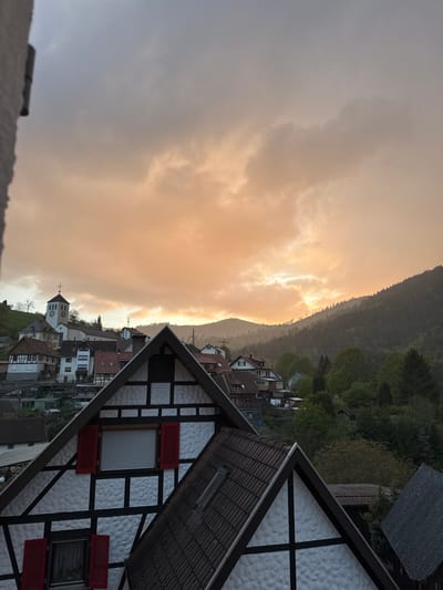 This image presents a picturesque, panoramic view of the town of Forbach, Germany, nestled at the base of a forested mountain range, likely captured during either sunrise or sunset. The sky, occupying the left side of the frame, is filled with dramatic clouds. A warm, golden light breaks through the clouds near the horizon, illuminating the mountain ridge and creating a vivid contrast with the muted gray tones of the clouds elsewhere.
A dense, dark green forest covers the mountain, which spans horizontally across the middle of the image, appearing as a dark silhouette against the glowing sky. Below the mountain, the charming town of Forbach unfolds, characterized by traditional German architecture. Numerous houses with reddish-brown tiled roofs dot the landscape. A prominent church, featuring a spire and a clock tower topped with a cross, stands out among the buildings towards the center-left of the town. Power lines are visible running across parts of the town.
In the immediate foreground, on the bottom right, is the gable end of a half-timbered house. Its white stucco walls are patterned with dark timber beams. This house features several windows, some adorned with bright red louvered shutters, and one with a closed white roller blind. The tiled roof of this foreground structure slopes downwards. The scene evokes a sense of peacefulness and atmospheric beauty in a historic German mountain village. No people are visible, and the time on the church clock face is not discernible.