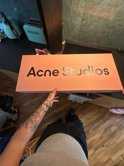 An overhead shot shows a person standing next to an "Acne Studios" shoebox. The box is salmon-colored with the name printed in alternating black and shimmering silver letters. The person's right hand is extended, pointing to the box, and their arm is tattooed and adorned with several bracelets. They are wearing black pants and a light-colored shirt. The setting is a domestic interior with a wooden floor, a countertop with kitchen appliances, and tiled walls. This is not enough information to determine a location in Saint-Ouen-sur-Seine, France.