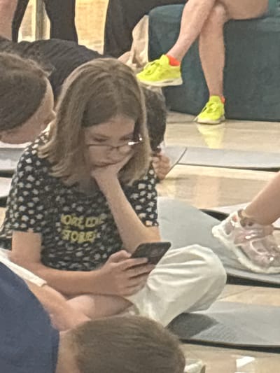 In an indoor setting, possibly a community center or a yoga studio in Tri Protoka, Russia, a young girl with blonde hair and glasses is sitting on the floor, leaning on her hand and looking at her mobile phone. She is wearing a dark-colored t-shirt with a white floral pattern and yellow text that reads "WORLD OF STORIES". Next to her, other people are partially visible, including someone in black pants and another person with light-colored athletic shoes. The floor appears to be tiled or polished concrete. The lighting is dim and diffused.