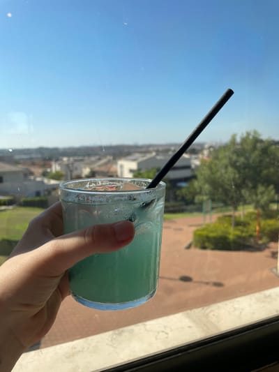 A hand holds a short, clear glass filled with a blue-green liquid, likely a cocktail, featuring a black straw. Condensation is visible on the glass. Outside the window, a cityscape stretches into the distance beneath a clear blue sky. A tree and portion of a brown-paved area are also visible. The photo seems to have been taken indoors.