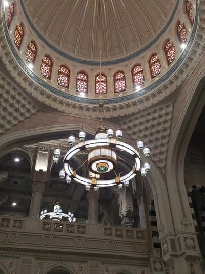 The image shows the interior of a building with a large dome and intricate architectural details, likely a mosque. A prominent circular chandelier hangs from the center of the dome. The dome features numerous small windows with decorative latticework. The walls below the dome are adorned with textured patterns and arched structures. Another, smaller chandelier can be seen in the background above a decorated balcony or mezzanine level. Given the architectural style and context, it is most likely a mosque in Makkah Al Mukarramah, Saudi Arabia. There are no people visible in the image.