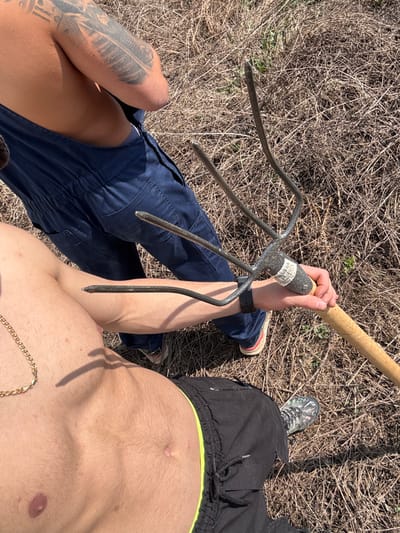 A top-down shot shows two individuals and a pitchfork against a background of dry grass. The person on the left wears navy overalls, sporting a prominent arm tattoo, while the other is shirtless with dark shorts featuring a neon yellow waistband and a gold chain around the neck. Both individuals are standing on the dry grass. The pitchfork is held in one hand, obscuring part of the overall-clad person's arm. One of the shirtless person's legs is visible as well, and they wear a shoe.
