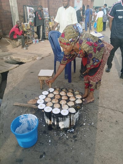 An African woman in colorful clothing sits on a blue chair cooking small, round, white patties on a charcoal grill. She is bent over the grill, seemingly monitoring the cooking process. A blue bucket sits on the ground nearby. The scene is outdoors, possibly a street or marketplace, with people and buildings in the background, including a storefront. The image suggests a street food vendor preparing a local dish.