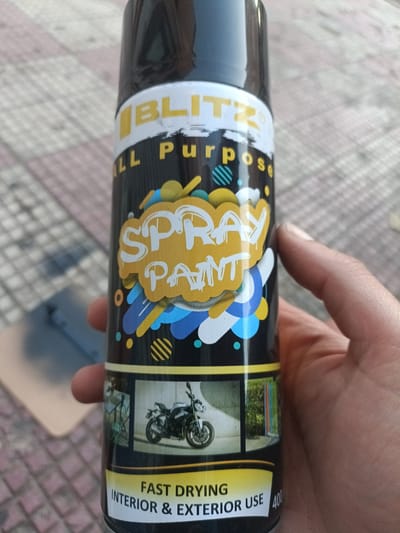 A person's hand holds a black spray can with colorful labeling, including "BLITZ ALL Purpose SPRAY PAINT," a graphic of a motorcycle, and text indicating "FAST DRYING INTERIOR & EXTERIOR USE." The background appears to be an outdoor tiled surface.