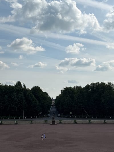 This is a full, high-angle shot of the Grand Menshikov Palace in Gatchina, Russia. The sky is a light blue with scattered white clouds. In the foreground, a wide, reddish-brown paved area stretches to the horizon. Two people, a woman in a dark top and light skirt and a man in a light blue outfit, are walking across this area. A stone wall with what appear to be cannons or decorative elements along its top separates this paved area from a street lined with dark green trees on either side. Cars are parked along the street. In the distance, at the end of the tree-lined avenue, a statue stands on a pedestal, and further beyond that, the Grand Menshikov Palace is visible with its distinctive dome and architecture.