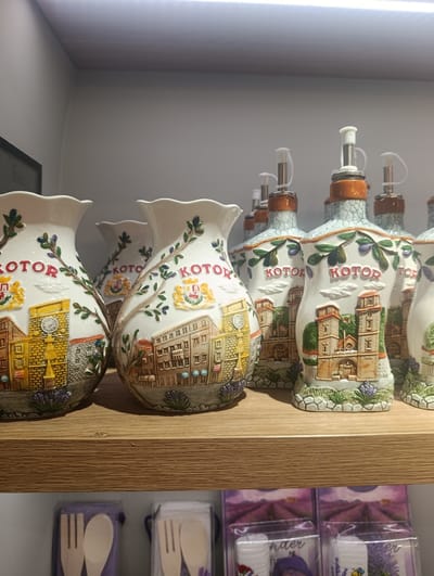 This image shows a shelf in a store displaying decorative ceramic items. The subjects are several vases and oil/vinegar dispensers adorned with detailed relief depictions of a European town, likely Kotor, Montenegro, given the visible text "KOTOR" in red lettering on some of the items. The town scenes feature buildings with windows, a clock tower on one vase, and what appears to be a coat of arms. Olive branches and sprigs of lavender are also integrated into the decorative designs.

The oil dispensers have silver spouts with white plastic caps and stoppers, indicating they are functional kitchenware designed for display. The vases have wide, scalloped rims and are also decorated with the town motif and similar natural elements.

The items are arranged on a light-colored wooden shelf, with other shelves visible above and below. The lighting appears to be artificial, coming from above and casting a slight glare on the top of the shelf. The overall impression is of a souvenir shop or a home decor store. No people are visible in the image. The lower shelf shows packaged items, possibly more souvenirs, with imagery of lavender fields. The focus is on the craftsmanship and detail of the ceramic collectibles.