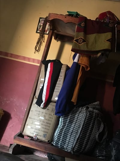 A cluttered wooden shelf with various items rests against a wall with a red and yellow color scheme. A woven, patterned vest, blue and yellow clothing, and a black and white striped item are stacked on top. The shelf also holds a picture frame with beads and sits on a box labeled "SON/MP 00912 FACE332" and other shipping details, including item and engine numbers. The box dimensions are listed as 355x330x480 mm.