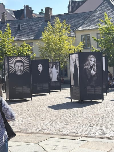 An outdoor photography exhibition is displayed on a cobblestone plaza in the city of Lund, Sweden. Several large, black-framed display panels feature black and white portraits. The first panel on the left shows a man with a beard and a radiant halo effect behind his head. To his right, a second panel displays a young woman with dark hair. Further to the right, a third panel shows a woman in a white cloak, and the panel to her right features a woman giving a middle finger gesture. Behind the displays, buildings with red-tiled roofs and light-colored facades are visible, surrounded by green trees. Sunlight casts distinct shadows on the plaza. A person in a striped shirt and jeans is partially visible in the foreground, carrying a black bag. Other individuals are present in the background, some seated and others standing.
