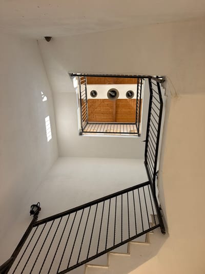 The image captures an interior view looking upwards from a staircase, within a building located in Kocherinovo, Bulgaria. The staircase has white steps and a dark, ornate metal railing. A black outdoor-style lamp is affixed to the wall near the railing. The walls are painted white, and a skylight with wooden slats and three circular fixtures is visible overhead. The overall impression is of a stairwell in an older building.