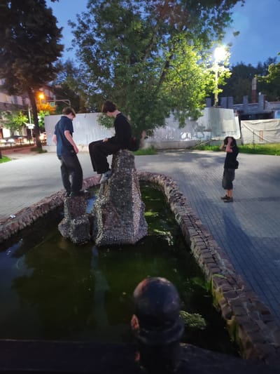 The image depicts a twilight scene in Shchyolkovo, Russia, featuring three young individuals near a decorative fountain. Two of the individuals, dressed in dark clothing, are positioned on or around a rock structure in the center of the fountain. One is seated on the peak of the structure, while the other stands beside it. The third individual, also in dark attire, stands to the right, facing away from the camera and appearing to adjust their hair or clothing.

The fountain itself has a stone border and contains murky green water with some visible debris. The surrounding ground is paved with interlocking bricks, and a tree with lush green foliage is prominent in the mid-ground, illuminated by a bright streetlight. In the background, there are construction barriers, suggesting ongoing development or renovation. Further back, a building with illuminated windows and streetlights are visible, indicating an urban or suburban environment.

The time of day is evident from the twilight sky, which is a deep blue, and the artificial lighting from the streetlamp. The weather appears calm, with no visible precipitation or strong winds. The overall mood conveyed is relaxed and casual, with the individuals seemingly engaged in spontaneous activity or posing. No discernible text is visible in the image.