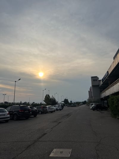 The image depicts a parking lot in front of a commercial building, possibly in San Martino Buon Albergo, Italy. The sun is visible in the cloudy sky, casting a soft glow. Several cars are parked in neat rows. The building appears to be multi-story with a modern facade, featuring signs indicating businesses. There are streetlights along the side of the parking lot and some trees and greenery are visible. The asphalt road surface shows some cracks. A few individuals are walking in the distance.