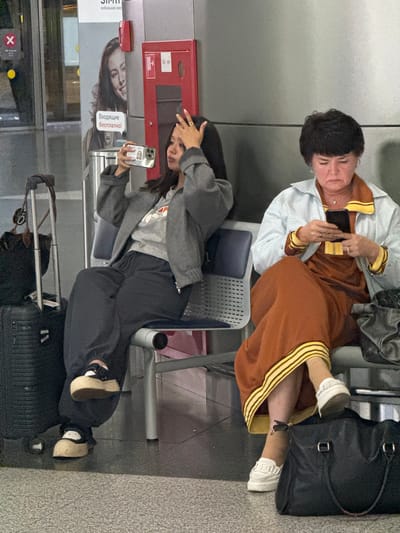 In Vnukovo, Russia, at an indoor location resembling an airport or train station waiting area, two women are seated. The woman on the left is younger and is taking a selfie with her phone, holding it in her left hand and touching her forehead with her right hand. She is wearing a gray jacket over a gray t-shirt and dark loose-fitting pants. She has on platform sneakers. Luggage, including a black suitcase and a brown bag, is visible beside her.
The woman on the right is older and is looking down at her phone, which she holds in her right hand. She is wearing a light blue jacket over an orange dress with yellow trim. She has on white sneakers. A large black duffel bag is on the floor next to her. The environment features a metallic wall with a red emergency box mounted on it and a poster with an image of a smiling woman. The seating consists of individual chairs with metal frames and blue padded seats. The floor is tiled.