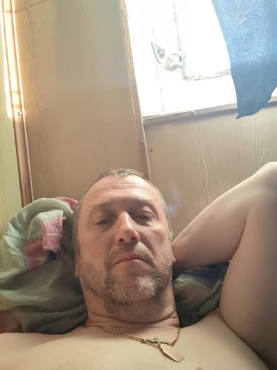A middle-aged Caucasian male is depicted in a selfie, taken from a low-angle perspective while he is lying down indoors in Ivanov Bor, Russia. He has short, curly, graying brown hair and a short, graying beard and mustache. His expression is neutral as he looks directly into the camera. He is bare-chested, wearing a gold-colored chain with a cross pendant and a rectangular tag around his neck. His head rests on a light green pillow with a pink patterned section. To his left, a partial view of his upper arm/shoulder is visible, with what appears to be a bent leg (likely his left) in the upper right portion of the frame. The background features light brown wooden wall panels. A window is visible in the upper right, admitting bright daylight, with a blue patterned curtain partially obscuring it. The window frame shows signs of wear and includes visible hardware.