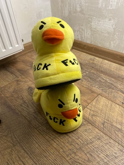 Two yellow duck-shaped slippers are stacked vertically on a wooden floor in an indoor environment. The slippers have orange beaks, black eyes, and black lettering that reads "FUCK" on the sides of the lower slipper and is partially visible on the upper slipper. To the left of the slippers, a portion of a white radiator is visible against a textured, light-colored wall. The floor is made of wooden planks.