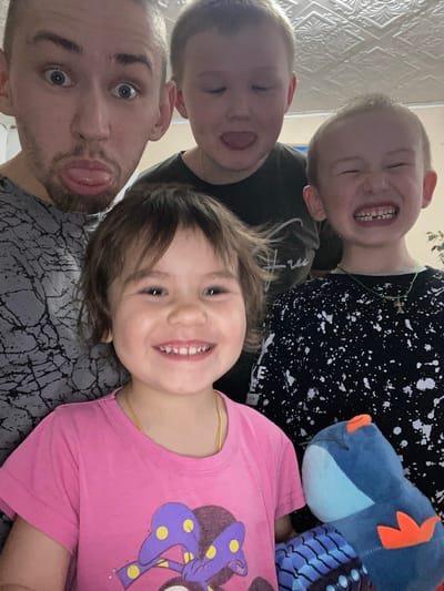 The image shows a selfie of a man with three children and a blue stuffed animal. The man is in the bottom left of the frame, sticking his tongue out. Above him is a child sticking their tongue out as well. To the right of these two is a young girl smiling at the camera. Above her is a child laughing, with missing front teeth. The children are wearing different patterned tops. The blue stuffed animal is near the top right corner. The background appears to be a textured wall, and part of a plant can be seen at the very top. The exact location within Bely Klyuch, Russia, is not discernible from the image.