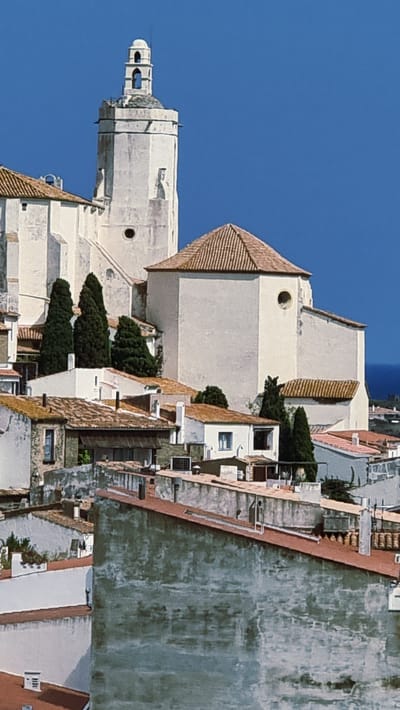 The image captures a view of the town of Cadaqués, Spain, dominated by white-washed buildings with terracotta roofs, clustered on a hillside under a deep blue sky. A prominent white church tower, with arched openings at its belfry and a dome-like cap, stands tall in the upper left quadrant of the image. Adjacent to the tower is a white, cubic structure with a pyramidal tiled roof and a circular window. The middle ground reveals a dense arrangement of smaller white houses with red tile roofs, interspersed with dark green cypress trees. In the foreground, more buildings with weathered, textured walls and red roofs are visible, creating a layered effect of architecture cascading down the slope. The distant horizon shows a sliver of the sea. There are no people or overt actions depicted in the scene; it is a static architectural and landscape composition.