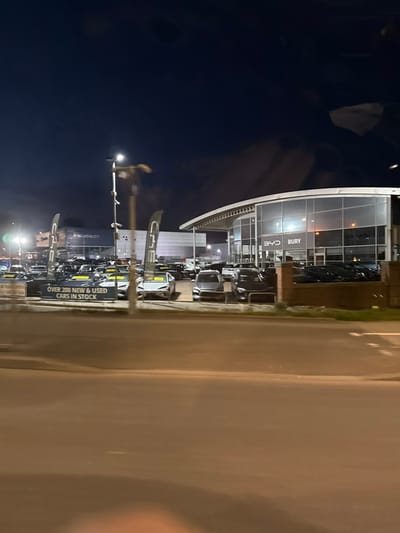 This nighttime image captures a commercial automotive sales area in Bury, United Kingdom. The scene is viewed from across a multi-lane asphalt road, which occupies the lower third of the frame. The dealerships are brightly illuminated by various artificial light sources.
On the left, a Peugeot dealership building is visible in the background. In the midground, a large inventory of new and used cars is parked, with a prominent sign stating "OVER 200 NEW & USED CARS IN STOCK." Several vertical white flags bearing a dark logo, possibly 'CME', are interspersed among the vehicles.
To the right, a modern, two-story car showroom features extensive glass paneling and a distinctive curved roof. The text "BYD BURY" is affixed to its facade, indicating a BYD dealership location. Additional vehicles are parked in front of this building, some partially obscured by a low brick wall. A tall street light with multiple luminaires stands centrally, casting light across the lot. The sky is dark, consistent with nighttime conditions. No individuals are visible in the frame.