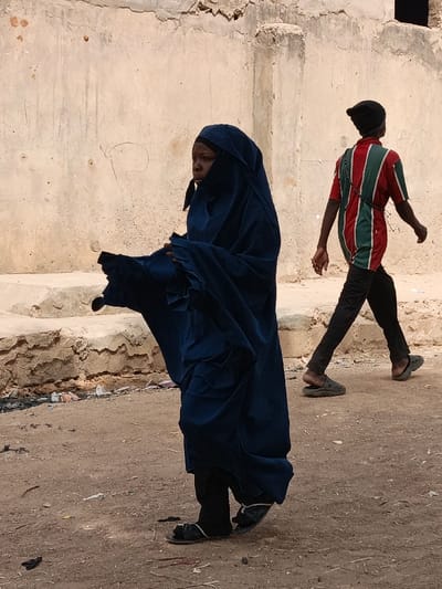 The image depicts a street scene in Zango, Nigeria, under bright daylight conditions. In the foreground, slightly to the left of center, an individual identified as an adult female stands, partially facing away from the viewer, with her head turned to the left. She wears a dark blue full-body covering, including a headscarf and face veil exposing her eyes and part of her lower face, over dark trousers and black open-toe sandals. Her left arm is slightly raised and extended forward with an open hand, suggesting a gesture or holding an unseen object, while the fabric of her covering billows around it. In the background, to the right, an adult male walks from left to right, his back mostly towards the camera. He wears a vertically striped red, green, and white short-sleeved t-shirt, dark trousers, and dark green/grey slip-on clogs, with his left heel partially out of the shoe. A dark beaded strap or necklace crosses his back from his left shoulder to his right hip, and white earbud wires are visible around his neck. He also wears a black beanie. The ground is an uneven, light brown dirt or sand surface with scattered debris. The background features a weathered, light beige wall with cracks, peeling plaster, and dark markings, consistent with an old building, above a lower, rough concrete or mud structure.