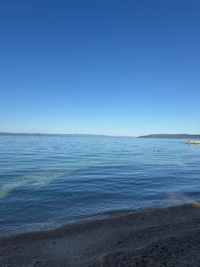 The image depicts a serene coastal scene in Makarska, Croatia. The vast expanse of the Adriatic Sea dominates the frame, its surface a tranquil blue under a clear, cloudless sky. The water is calm, with gentle ripples reflecting the sunlight. In the foreground, a pebbly beach lines the edge of the water. The beach is sparsely populated, with a few small buoys marking an area in the sea. In the distance, a hazy mountain range forms the horizon, characteristic of the Makarska Riviera. A small rocky outcrop is visible on the right side of the frame, with a couple of people seated on it, enjoying the view. The overall impression is one of peace and natural beauty.