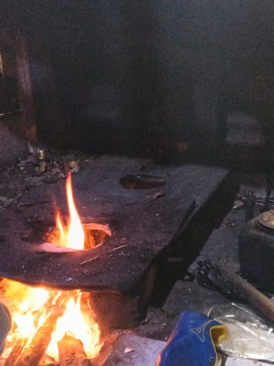 This image, taken in Na Sangkham, Laos, depicts a low-light scene centered on a traditional stove. In the lower left quadrant, a vigorous fire with orange and yellow flames burns within the main combustion chamber of a dark, stone or concrete stove. Additional flames emanate vertically from a circular opening on the top surface of the stove. Another circular indentation is present on the stove's surface further to the right. Wood fragments serve as fuel for the fire. To the right of the stove, a dark, lidded pot or kettle rests on a dark surface. A long, dark object, possibly a tool or structural component, extends diagonally from the lower right toward the center. In the immediate lower right foreground, a blue glove with yellow trim is visible, resting on a reflective surface, possibly plastic sheeting. The immediate ground area around the stove contains scattered debris. The background is predominantly dark and indistinct due to the low ambient light, with illumination primarily provided by the fire. No individuals are visible in the frame.