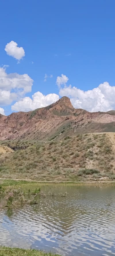 The image depicts a natural landscape dominated by a mountain range under a bright blue sky with scattered white clouds. A body of water, likely a lake or pond, occupies the foreground, reflecting the sky and surrounding terrain. The mountains are rugged and appear to be composed of reddish-brown rock, with patches of green vegetation on their slopes. The foreground features dry, grassy terrain leading to the water's edge, with some sparse vegetation growing in and around the water. The overall impression is one of a serene and undeveloped natural environment. The location is identified as Lanjazat, Armenia. There are no people or animals visible in the image, and no human-made objects are prominently featured, suggesting a focus on the natural beauty of the landscape.