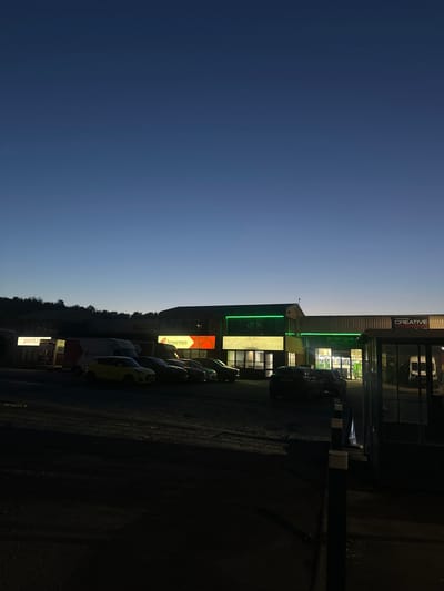 This vertically oriented image captures a commercial or retail park at dusk or early night in Tonbridge and Malling, United Kingdom. The upper portion of the frame features a dark blue, clear sky, with a faint crescent moon visible in the upper left.
The right side of the image (top if rotated correctly) is dominated by several low-rise commercial buildings, brightly illuminated by their signs. Visible businesses include "CREATIVE DESIGNS" and "énergie fitness", with the latter displaying green neon signage that also advertises "ONLY £23.99". Another prominent business is "Bournes", identified by a red and white arrow sign on a building and matching branding on a white parked van. A partial sign reading "practi" is also visible on one of the buildings.
A paved parking lot occupies the foreground and mid-ground, containing several parked vehicles, including a yellow car and the white Bournes van. Dark silhouettes of trees are visible behind the buildings in the distance. The scene is static, showing no active events or people, and is characterized by the contrast between the darkening natural light and the bright artificial illumination of the business signs.