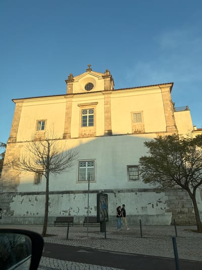 This image depicts the exterior of the Igreja do Seminário de Santarém, also known as the Church of Our Lady of Piety, located in Santarém, Portugal. The perspective is a low-angle shot from street level, likely from a vehicle, with a blurred side mirror visible in the bottom-left foreground.
The prominent structure is a large, multi-story historic building with an off-white/pale yellow stucco facade. The upper portion of the facade is brightly illuminated by direct sunlight, while the lower portion is partially in shadow. The building features classical architectural elements, including stone pilasters, a prominent cornice, and a central pediment topped with a stone cross. A circular oculus window is set within the pediment. Multiple rectangular windows of varying sizes are present across the facade; some have white frames and multiple panes, while others display dark grilles. Traditional terracotta barrel roof tiles are visible along the building's top edge. The lower half of the facade exhibits significant wear, with peeling stucco revealing underlying stone blocks and rough plasterwork.
The foreground consists of a cobblestone pavement with a narrow strip of asphalt in the immediate foreground. Several dark metal bollards delineate the edge of the paved area. Two dark brown wooden park benches are situated on the sidewalk. A black advertising billboard, partially displaying "IKEA" text, stands on the left side of the sidewalk.
Two individuals are walking from left to right on the cobblestone sidewalk in the mid-ground. The person on the right is male, wearing dark trousers, a dark long-sleeved shirt, and sunglasses. The person on the left is female, wearing light blue jeans and a dark short-sleeved top. They are walking side-by-side.
Two trees are present: a leafless deciduous tree with bare branches stands in the left-center, casting a shadow onto the building's lower wall. A second tree with green foliage is positioned on the right, partially obscuring a window on the building. The sky is clear and deep blue, occupying the upper half of the frame. The strong directional light and distinct shadows indicate either early morning or late afternoon.