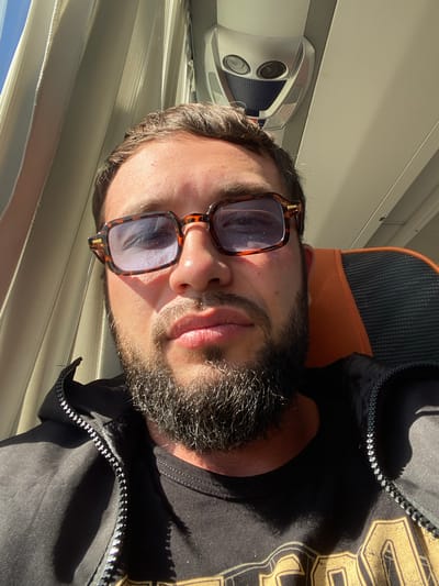 A man with a beard and sunglasses is taking a selfie in what appears to be a vehicle, possibly a bus or train, given the overhead compartments and window blinds. The man is wearing a black jacket zipped over a black t-shirt with a graphic design. Sunlight streams in from the left, casting shadows. The background shows interior elements of the vehicle, including seating and overhead fixtures. The exact location in the city of Терволово, Russia, is not determinable from the image alone.
