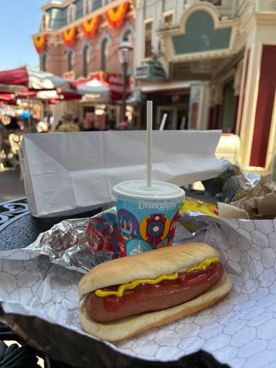A hot dog with ketchup and yellow mustard in a bun, resting on white hexagonal-patterned paper, occupies the foreground on a black ornate table. To its left, a piece of crumpled aluminum foil is visible. A light blue plastic cup with a white lid and straw, featuring "Disneyland Resort" branding and colorful geometric patterns, sits behind the hot dog. Further back, a white paper bag stands upright, with additional crumpled paper and packaging to its right. The scene is set outdoors in bright daylight. The background, largely out of focus, reveals a street within Disneyland Park in Anaheim, California, United States. Victorian-style buildings are visible, including a prominent light pink structure adorned with red and yellow scalloped banners, arched windows, and a dark roof. Red and white striped umbrellas cover outdoor seating areas, and blurred figures of people are present. Multiple "Coca-Cola" signs are discernible on distant buildings, along with a lamppost.