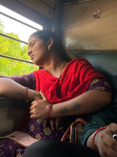 This media file shows a close-up of a woman lying down, possibly sleeping or resting, inside a train carriage. She is wearing a red garment draped over her shoulders and a purple patterned outfit underneath. Her head is tilted, and her eyes are closed. She has several bracelets on her wrists, including red and gold bangles. A tattoo of what appears to be an elephant is visible on her forearm.

The background shows the interior of a train, with metal bars of a window and a glimpse of green foliage outside, suggesting a daytime scene. Two small rectangular tags with the number "65" are visible on a metal surface in the upper left portion of the image. The overall lighting suggests it is daytime, and the greenery outside implies an outdoor, possibly rural, setting. The image is oriented vertically, with the subjects appearing upside down relative to a typical upright orientation.