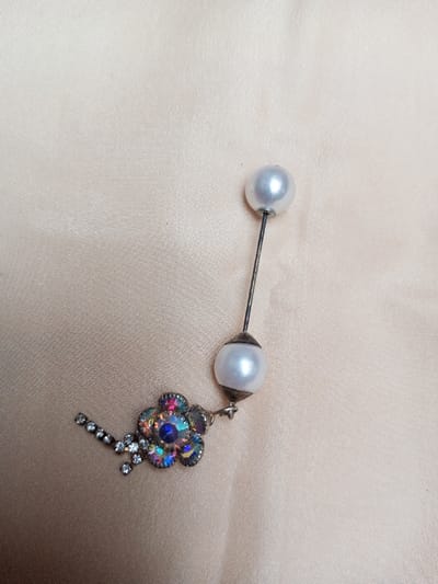 A decorative pin and attached charm are centrally positioned on a light beige, subtly reflective fabric surface, possibly satin, exhibiting minor creases. The pin consists of a slender, dark metallic rod, approximately 3-4 cm in length, terminating at both ends with spherical, white, lustrous beads, resembling pearls, each approximately 8-10 mm in diameter. The metal rod appears slightly tarnished. A decorative element is suspended from the lower pearl bead via a small metallic ring. This element comprises a multi-faceted, flower-shaped component with iridescent, possibly aurora borealis-coated, central and petal-forming stones, and an extension featuring a linear arrangement of small, clear, faceted rhinestones. The metallic settings for the stones appear silver-toned. The scene is located in Qarabucaq qəsəbə, Azerbaijan.