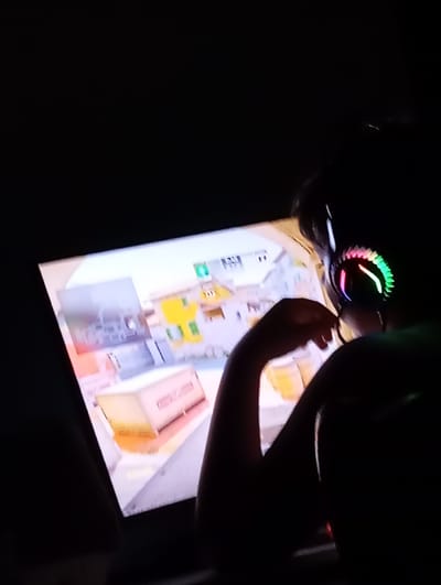 A person with headphones is playing a video game on a screen in a dark room. The video game appears to be a first-person shooter, and the screen shows a building with yellow walls and boxes. The headphones have rainbow-colored lights. The person's hand is visible in front of the screen. The location in the city of Yukariyuz, Uzbekistan is not apparent from the visual information provided.