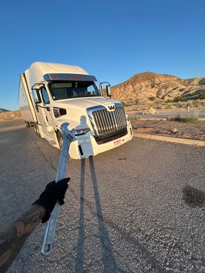 A first-person perspective shot shows a gloved hand holding a large adjustable wrench in front of a white semi-truck parked on an asphalt road. The truck's grille is prominent, with the "W" logo visible. The truck has a long trailer attached. In the background, arid, rolling hills with sparse vegetation are visible under a clear blue sky. The sun is low, casting long shadows. The scene appears to be a rest stop or pull-off area in a desert landscape. There is no indication that the location is in Salina, United States of America.
