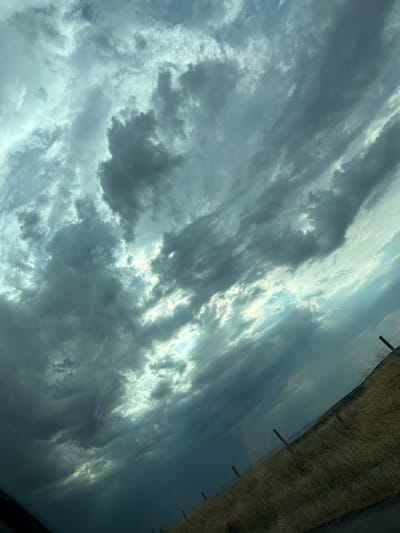 The image shows a sky filled with dark, dramatic clouds, suggesting an impending storm. Sunlight breaks through in some areas, creating a contrast between light and shadow. The lower portion of the image features a dry, grassy field with a wire fence. The scene is tilted, indicating it was likely taken from a vehicle. The location of Zheleznik, Bulgaria, is not discernible from the visual information presented in the image.