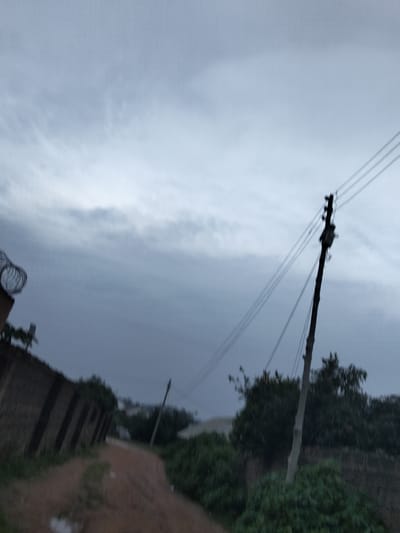 The image depicts an overcast sky over a rural or semi-rural landscape. A dirt road, wet in places, curves through the scene. To the left, a brick wall topped with barbed wire runs along the road. On the right side of the road, a wooden utility pole supports overhead wires, and trees and dense foliage are visible. In the distance, more trees and some structures can be seen under the gloomy sky. The overall impression is one of a somber or approaching storm atmosphere. The location is identified as Barikin Jirga Summa, Nigeria.