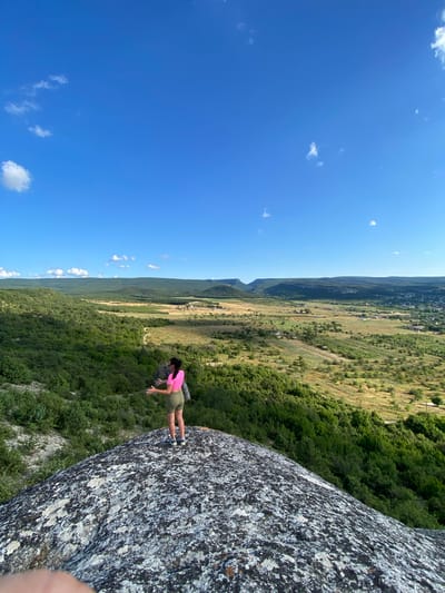 A woman stands on a large boulder, looking out over a vast, green valley under a bright blue sky with scattered white clouds. She is wearing a pink crop top and olive green shorts, with her back to the camera and her arms outstretched. The boulder is speckled with white and grey lichen. The valley below is covered in lush green trees and shrubs, with patches of yellowed grass and a small village nestled in the distance. Rolling hills and mountains form the horizon. The scene appears to be set in a natural, rural landscape within the Krasnomakskoye rural settlement of Ukraine.
