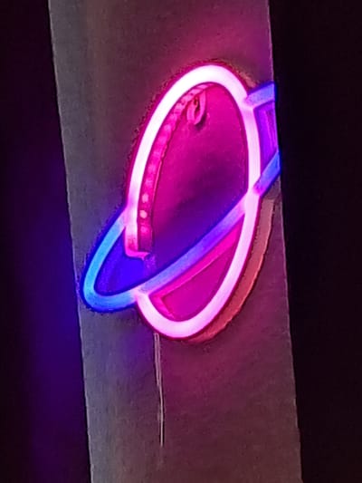 The image displays a neon light fixture shaped like a planet with rings, affixed to a vertical surface that appears to be a pillar or wall. The neon colors are predominantly pink and blue, with the planet itself glowing with a bright pink and white light, and the rings emitting a blue and pink glow. The surface the fixture is attached to is textured and gray, contrasting with the vibrant illumination. The background is dark and out of focus, suggesting a dimly lit environment. There is a thin, white wire extending downwards from the fixture. The provided context states this is in Parchim, Germany, but no specific location within the city is discernible from the image alone. There are no people, actions, or interactions depicted.
