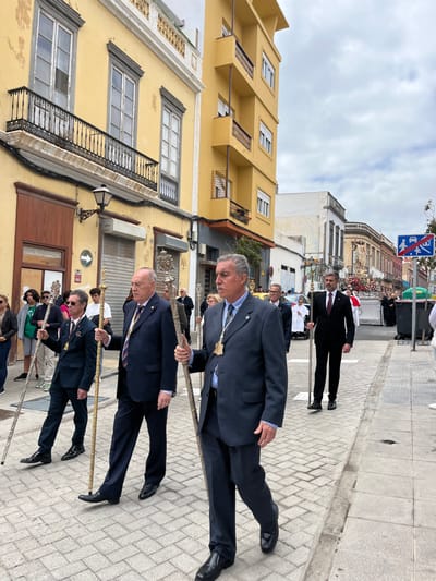 The image shows a procession of men walking down a street in the Vegueta district of Las Palmas de Gran Canaria, Spain. The men are wearing suits and carrying staffs. There are buildings on both sides of the street, and the pavement is made of grey brick. The men are walking in a single file line, and they appear to be participating in a religious procession. The sky is overcast with a light grey color. The sun is not visible. The time is likely midday or early afternoon.
