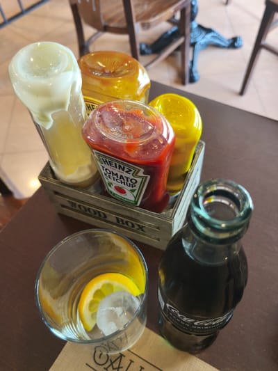 The image presents a close-up view of a casual dining table setting, likely in a restaurant or cafe in Pontevedra, Spain. In the center, a rustic wooden caddy, labeled "WOOD BOX," holds three inverted squeeze bottles of condiments. These include a bottle of mayonnaise with its creamy contents visible, a prominent Heinz Tomato Ketchup bottle, and a bright yellow bottle of mustard.
To the right of the condiment caddy is a classic glass bottle of Coca-Cola. To the left, a clear drinking glass is prepared with ice cubes and a slice of lemon, ready for a refreshing beverage. The table surface is a dark wooden material.
The background is softly blurred, revealing parts of wooden chairs and a light-colored tiled floor, indicating an indoor setting. The ambient lighting suggests it is daytime. The setup implies an imminent meal or a break at a casual eatery.
Visible text includes "WOOD BOX" on the condiment caddy. The ketchup bottle clearly shows "HEINZ", "EST 1869", "TOMATO KETCHUP", and partially "100% S..." (likely 'SQUEEZE'). The Coca-Cola bottle's label reads "Coca-Cola" and "SABOR ORIGINAL." A partial logo, "OXL...", is visible on the paper napkin or placemat beneath the drinking glass.