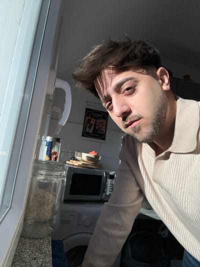 The image depicts a male individual in a domestic kitchen environment located in Remscheid, Germany. The subject, an adult male with dark hair and a trimmed beard, is positioned in the foreground, looking directly at the camera. He wears a light beige, ribbed polo shirt. Strong, direct sunlight enters from the left, illuminating the right side of his face and body, and casting distinct shadows across his forehead and the left side of his face. To the left, a white window frame with a matching handle is visible, bordering a speckled gray countertop. On this countertop, a large clear glass jar, appearing to contain a light-colored granular substance, and a smaller metallic container are present. In the background, kitchen appliances include a silver-colored microwave oven situated on a surface, above which a white bowl containing an orange substance rests on a small wooden base. A dark poster or framed picture is affixed to the wall, displaying text that includes "MY FAVORITE RECIPES" and "Enjoy Eating." Below the microwave, the upper portion of a white washing machine is visible.