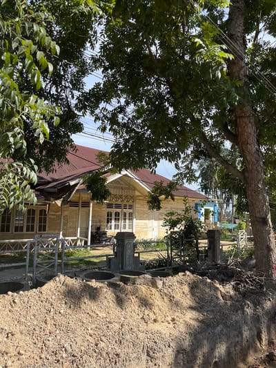 The image depicts a street scene in Kota Kuala Simpang, Indonesia, under a bright sky. In the foreground, a pile of excavated dirt and rocks sits prominently, suggesting ongoing construction or landscaping work. Beyond the dirt mound, a low white fence with a crisscross pattern borders a grassy area. Behind the fence, a light-colored, wooden building with a dark red roof is visible. The building features multiple windows with panes and a covered porch area. A white door is centrally located on the visible facade. In front of the building and to the right of the door, a dark-colored wheeled cart or vehicle is parked. A large tree with textured bark dominates the right side of the frame, its branches extending over the scene and partially obscuring the sky and overhead power lines. To the left, another tree with green foliage shades the building and the street. Further in the background, more trees and parts of other buildings are discernible, including a small structure painted blue with white trim. The overall impression is one of a tropical or subtropical environment with a mix of residential structures and natural elements.
