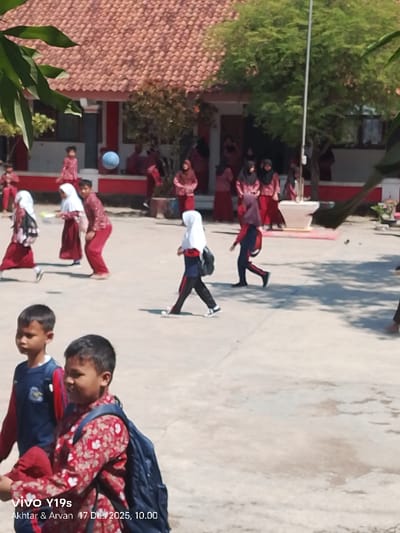 A group of children in school uniforms are visible in a schoolyard. Some are walking, others are standing near a red and white building. A blue ball floats in the air. Two young boys in the foreground wear backpacks; one is in a red patterned shirt and the other in a blue and red jacket. The timestamp "Akhtar & Arvan 17 Des 2025, 10.00" is visible at the bottom left corner.