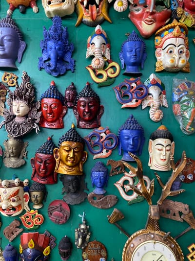 The image shows a wall filled with numerous colorful masks and decorative items, likely displayed for sale in a shop in Kathmandu, Nepal. The masks are of various sizes and depict faces of deities, animals, and symbolic figures, such as Buddha, Ganesh, and fierce demons. Many of the masks are made of wood and intricately carved and painted. Some of the masks are in the shape of heads, while others are flat wall hangings. Among the decorative items are Om symbols, a wooden clock with deer antlers, and carved wooden animals. The overall impression is of a vibrant and culturally rich display of traditional Nepali artistry.